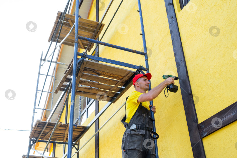 Скачать Мастер на все руки устанавливает деревянные балки на желтом фасаде, стиль фахверк, внешняя отделка дома, частный проект облицовки, ремонтные работы, благоустройство дома. фотосток Ozero