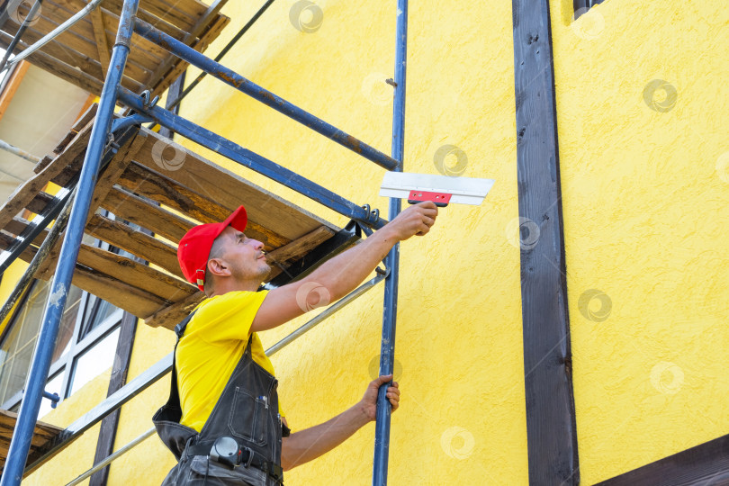 Скачать Декоратор с мастерком на строительных лесах, нанесение декоративной штукатурки, выравнивание фасадов, ремонт стен, частный бизнес, услуги по подготовке фасада здания. фотосток Ozero