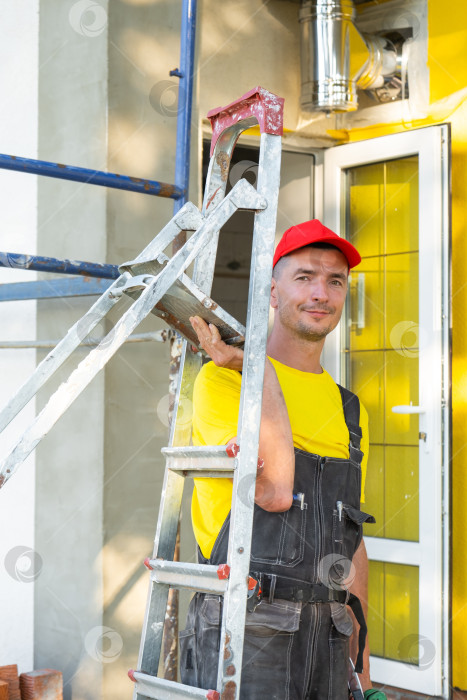 Скачать Портрет улыбающегося строителя со стремянкой возле дома, ремонт и благоустройство дома, компания по оказанию строительных услуг на месте, надежный эксперт. фотосток Ozero