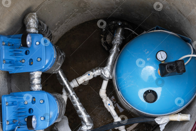 Скачать Установка системы внутри артезианской скважины: синий гидротанк, встроенные фильтры для воды, двигатель насоса, надежный источник воды для дома, установка и подключение насосного оборудования. фотосток Ozero