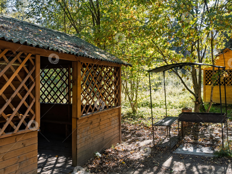 Скачать Деревянная садовая беседка с решетчатыми панелями и индивидуальной мангальной площадкой в тенистом осеннем парке фотосток Ozero