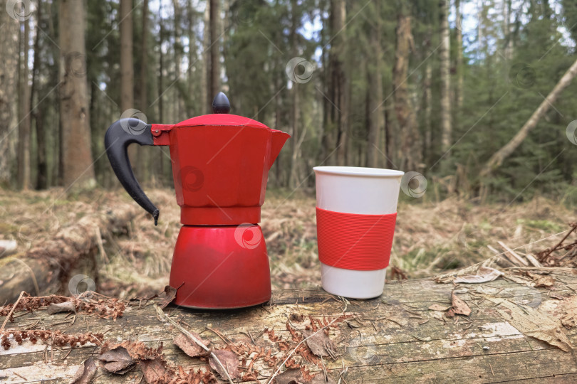 Скачать Красная кофеварка крупным планом стоит рядом с чашкой на покрытой мхом лесной подстилке. фотосток Ozero