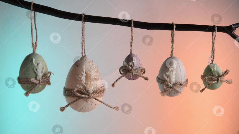Скачать Пасхальные яйца пастельных оттенков зеленого, белого и фиолетового цветов свисают с естественной ветки, перевязанные бантиками из бечевки и сухими листьями, на ярком фоне весеннего праздника фотосток Ozero