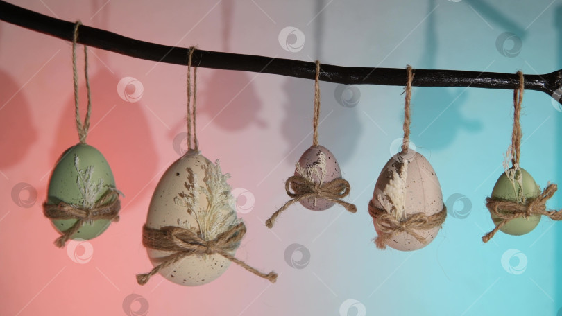 Скачать Пасхальные яйца пастельных оттенков зеленого, белого и фиолетового цветов свисают с естественной ветки, перевязанные бантиками из бечевки и сухими листьями, на ярком фоне весеннего праздника фотосток Ozero