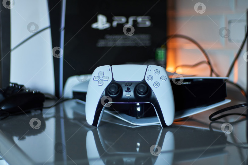 Скачать Тюмень, Россия - 8 июля 2025 года: Элегантная игровая консоль Sony PlayStation 5 установлена на современной поверхности и дополнена стильным контроллером, подключенным для зарядки фотосток Ozero