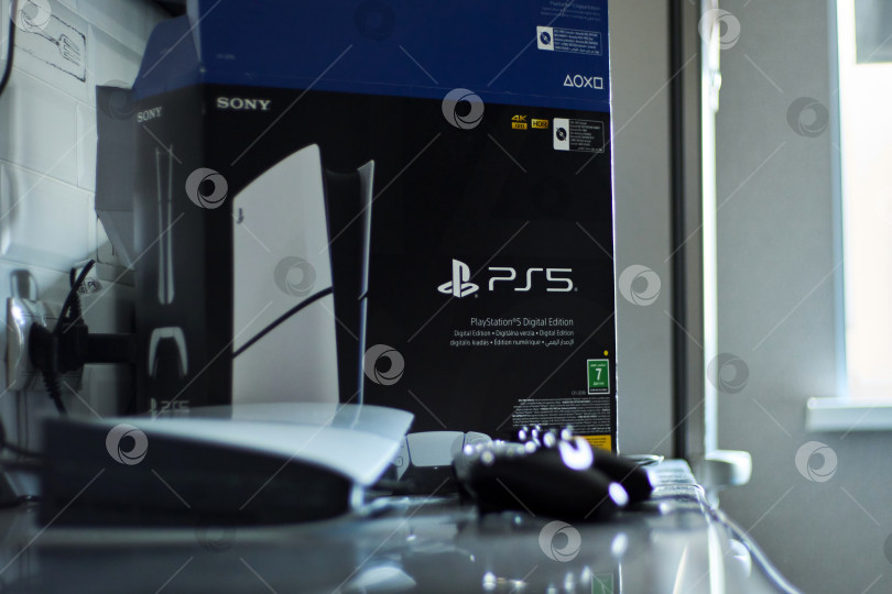 Скачать Тюмень, Россия - 8 июля 2025 года: Элегантная игровая консоль Sony PlayStation 5 установлена на современной поверхности и дополнена стильным контроллером, подключенным для зарядки фотосток Ozero