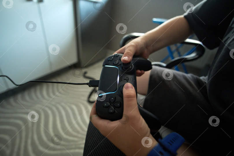 Скачать Тюмень, Россия - 8 июля 2025 года: Пользователь Sony PlayStation 5 работает с игровым контроллером, сидя в современной комнате в дневное время фотосток Ozero