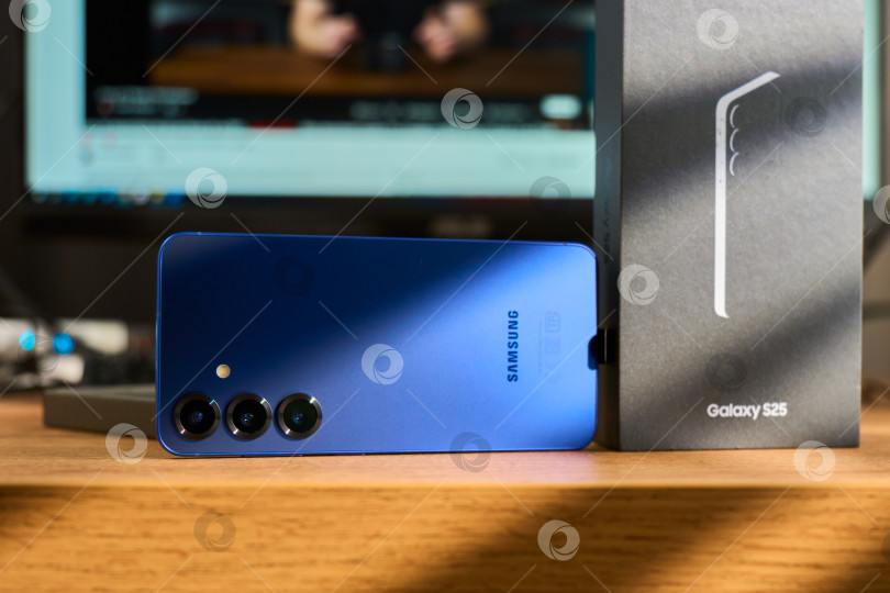 Скачать Тюмень, Россия - 3 декабря 2025 года: смартфон Samsung Galaxy S25 на деревянной поверхности рядом с коробкой, демонстрирующей детали технологии. фотосток Ozero