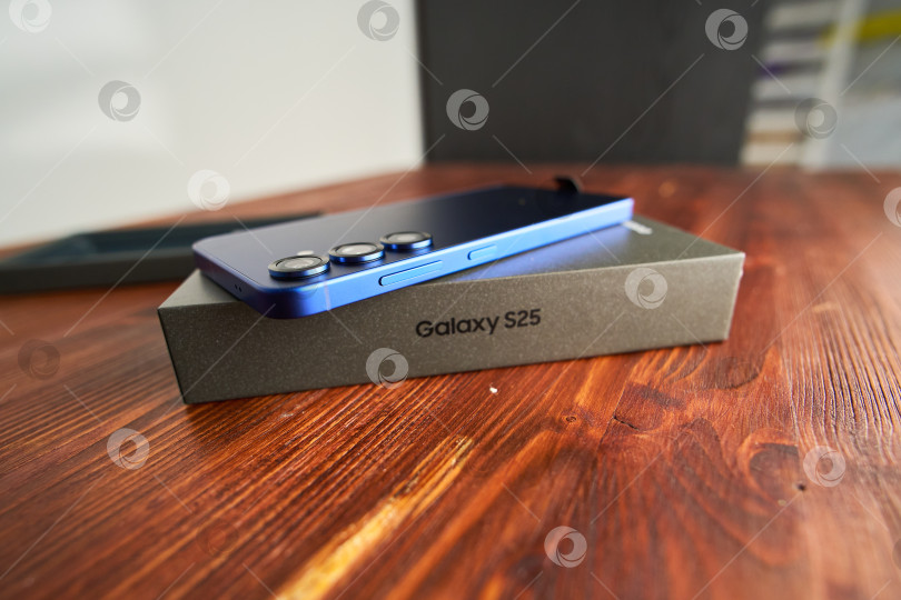 Скачать Тюмень, Россия - 3 декабря 2025 года: смартфон Samsung модели S25 в руках с упаковкой на деревянной поверхности. фотосток Ozero