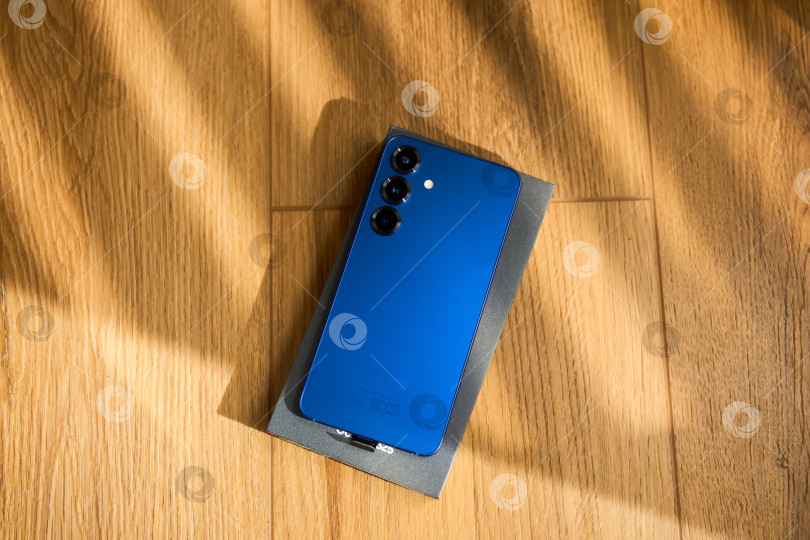 Скачать Тюмень, Россия - 3 декабря 2025 года: Новый смартфон samsung s25 стоит рядом со своей коробкой на деревянной поверхности, на которой четко видны основные детали фотосток Ozero
