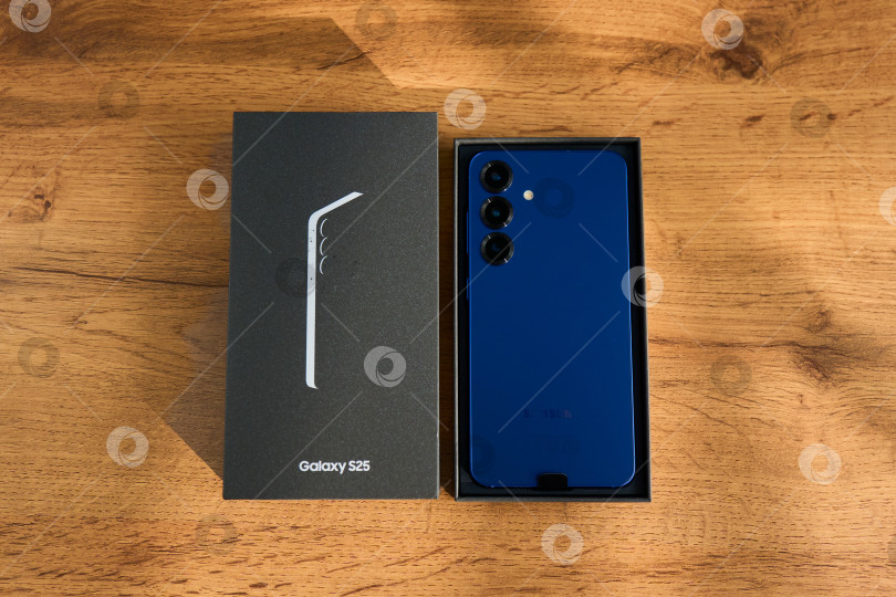 Скачать Тюмень, Россия - 3 декабря 2025 года: смартфон Samsung Galaxy S25 на деревянной поверхности рядом с коробкой, демонстрирующей детали технологии. фотосток Ozero