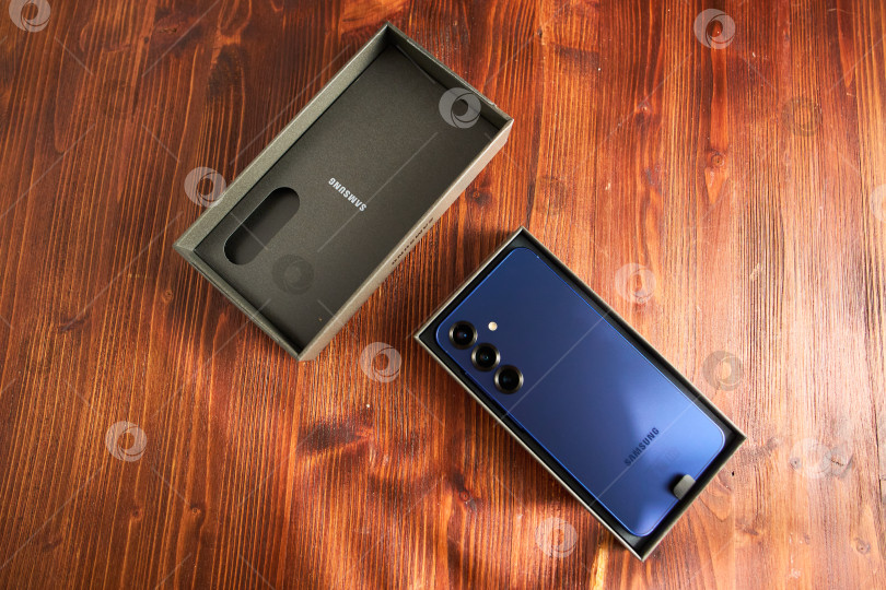 Скачать Тюмень, Россия - 3 декабря 2025 года: Новая модель смартфона Samsung s25 в коробке на деревянном столе с акцентом на функции камеры и фирменные детали. фотосток Ozero