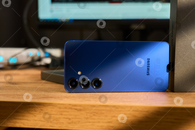 Скачать Тюмень, Россия - 3 декабря 2025 года: смартфон Samsung Galaxy S25 на деревянной поверхности рядом с коробкой, демонстрирующей детали технологии. фотосток Ozero