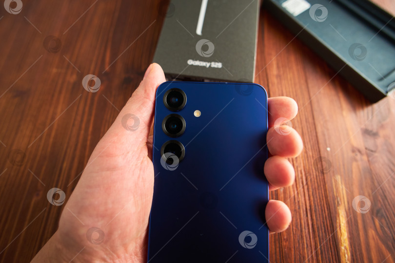 Скачать Тюмень, Россия - 3 декабря 2025 года: смартфон Samsung модели S25 в руках с упаковкой на деревянной поверхности. фотосток Ozero