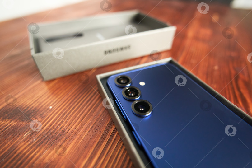 Скачать Тюмень, Россия - 3 декабря 2025 года: Новая модель смартфона Samsung s25 в коробке на деревянном столе с акцентом на функции камеры и фирменные детали. фотосток Ozero
