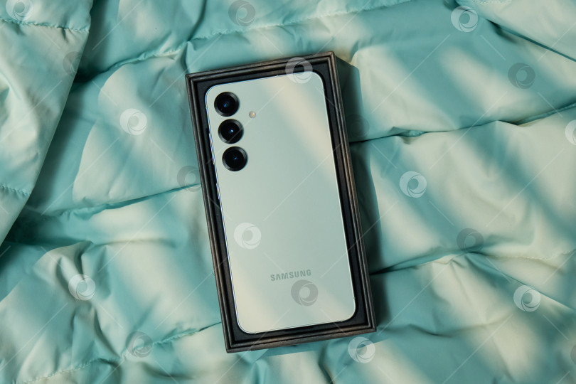 Скачать Тюмень, Россия - 11 декабря 2025 г.: Демонстрация устройств Samsung S25 на светло-голубом покрывале с мягким естественным освещением в непринужденной обстановке в помещении фотосток Ozero