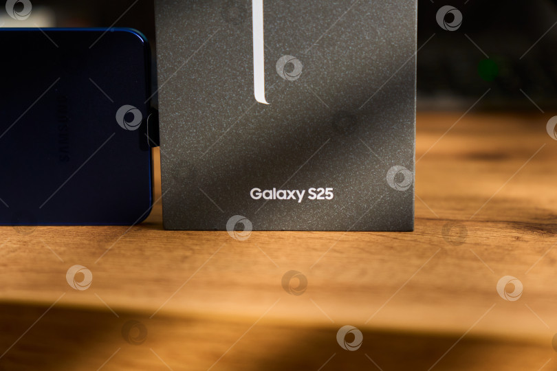 Скачать Тюмень, Россия - 3 декабря 2025 года: смартфон Samsung Galaxy S25 на деревянной поверхности рядом с коробкой, демонстрирующей детали технологии. фотосток Ozero