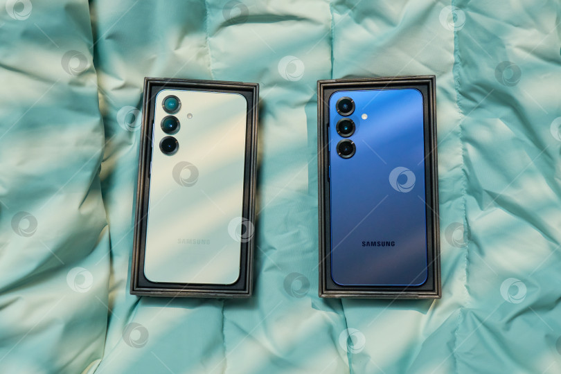 Скачать Тюмень, Россия - 11 декабря 2025 г.: Демонстрация устройств Samsung S25 на светло-голубом покрывале с мягким естественным освещением в непринужденной обстановке в помещении фотосток Ozero