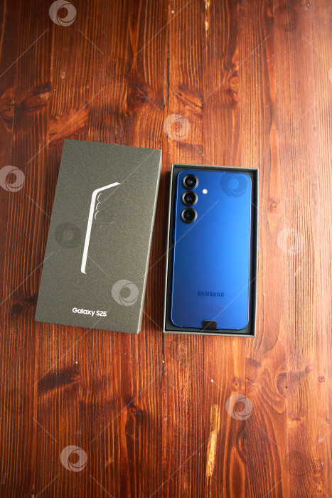 Скачать Тюмень, Россия - 3 декабря 2025 года: смартфон Samsung модели S25 в руках с упаковкой на деревянной поверхности. фотосток Ozero