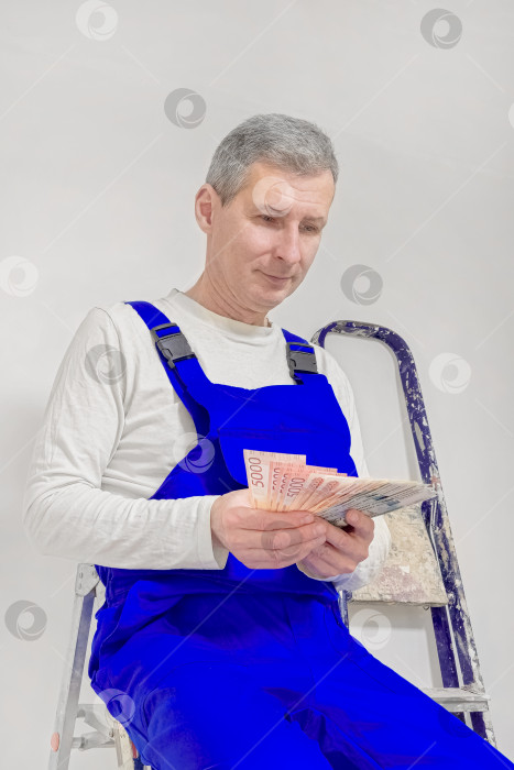 Скачать Вид сверху: штукатур сидит на лестнице, держа в руках российские деньги. фотосток Ozero