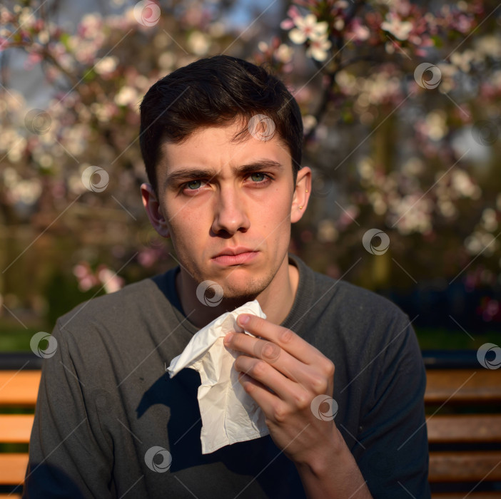 Скачать Молодой задумчивый мужчина, вытирающий нос салфеткой на фоне цветущих деревьев фотосток Ozero