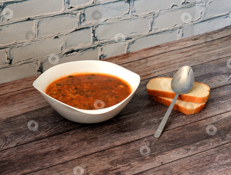 Скачать Острый кавказский суп харчо в глубокой тарелке на деревянном столе, рядом с ним ложка и два кусочка пшеничного хлеба. фотосток Ozero