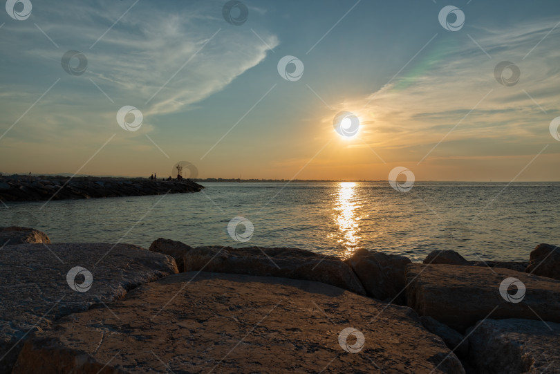 Скачать Закат над водой со скалами на переднем плане и людьми, прогуливающимися по берегу в прибрежной зоне фотосток Ozero