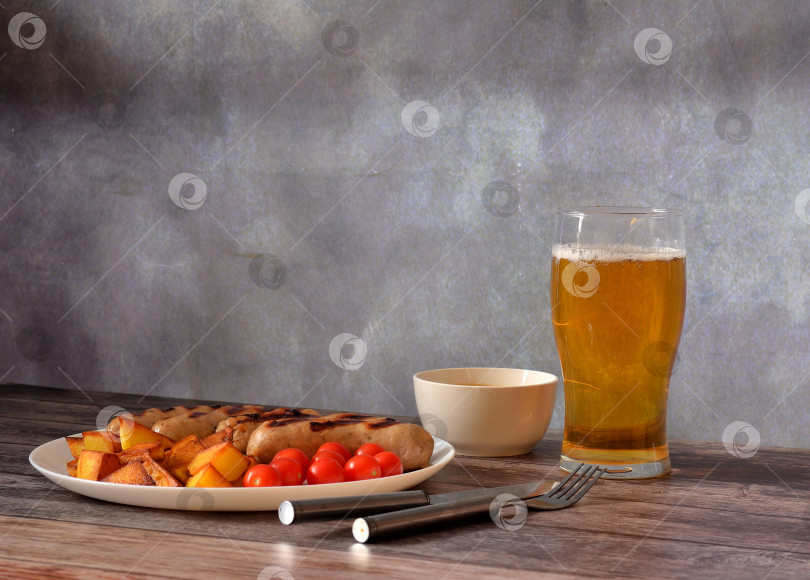 Скачать Бокал светлого пива, тарелка с жареными сосисками, отварным картофелем и помидорами черри на деревянном столе, рядом с чашкой соуса и столовыми приборами. фотосток Ozero
