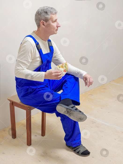 Скачать Рабочий в синем комбинезоне сидит на табурете. Он ест пиццу. Перерыв в работе фотосток Ozero