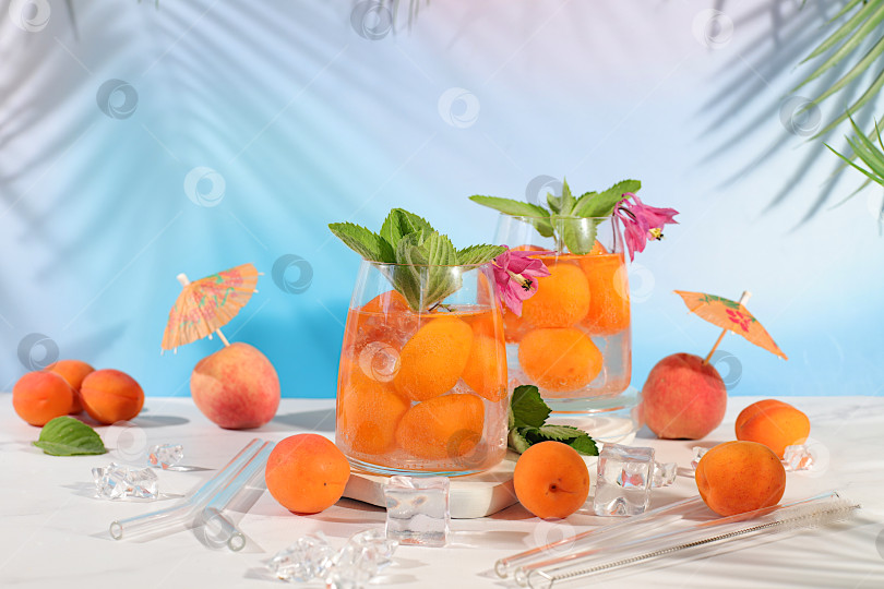 Скачать Персиковый чай со льдом или коктейль с мятой и абрикосами, рецепт освежающего летнего напитка. Два бокала с персиковым лимонадом на легком столе, концепция детокс-диеты и похудения, меню ресторана, фотосток Ozero