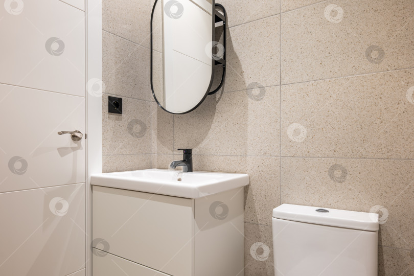 Скачать Компактная ванная комната с раковиной, краном, зеркалом и унитазом на фоне бежевой плитки с двойной дверью. Продуманная, но элегантная и простая концепция дизайна фотосток Ozero