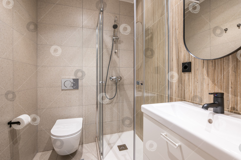 Скачать Небольшая стильно оформленная ванная комната со стильным зеркалом, белой керамической раковиной, туалетом и душем, отделанная бежевой плиткой. Концепция ванной комнаты в компактных домах или отелях фотосток Ozero