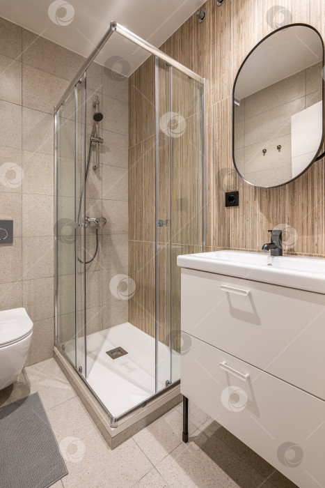 Скачать Диагональный снимок небольшой ванной комнаты со стильным белым туалетным столиком, стеклянной душевой кабиной и керамической раковиной, облицованной красивой бежевой плиткой. Концептуальная ванная комната после ремонта в квартире фотосток Ozero