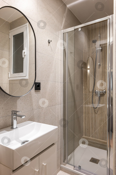 Скачать Удобная компактная ванная комната, отделанная стильной бежевой плиткой, со стеклянной душевой кабиной, раковиной и зеркалом современной формы. Концепция многофункциональной ванной комнаты в компактной квартире фотосток Ozero
