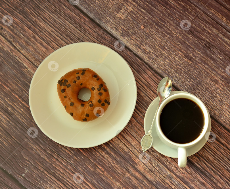 Скачать Свежий пончик с карамельной глазурью и топпингом на круглой тарелке и чашка горячего черного кофе на деревянном столе. фотосток Ozero