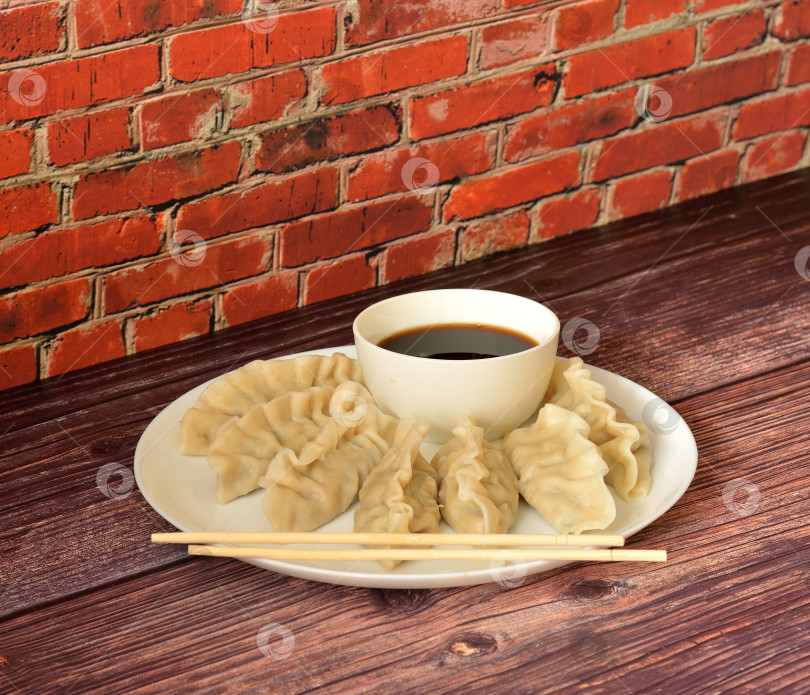 Скачать Тарелка свежих китайских пельменей под названием Вантоны с соевым соусом и двумя бамбуковыми палочками для еды на деревянном столе. фотосток Ozero