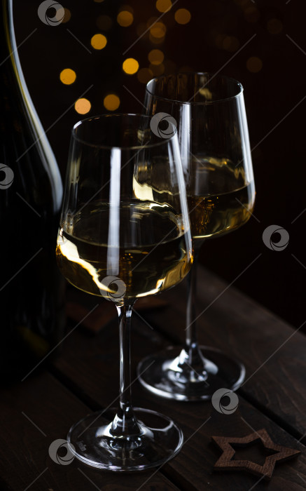 Скачать Два бокала белого вина и бутылка на деревянном столе, а за ними гирлянды огней. фотосток Ozero
