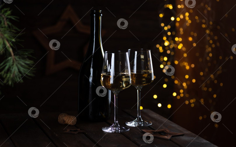 Скачать Два бокала белого вина и бутылка на деревянном столе, а за ними гирлянды огней. фотосток Ozero