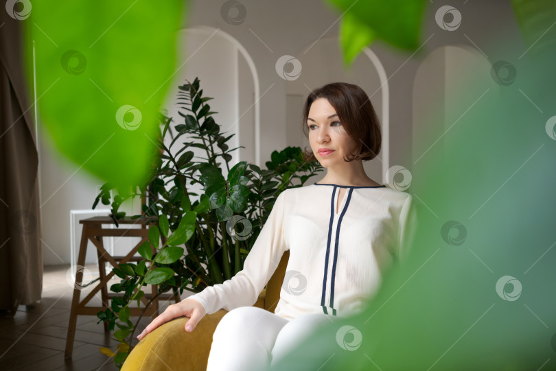 Скачать Безмятежная женщина в желтом кресле, смотрящая в сторону в биофильном интерьере с комнатными растениями фотосток Ozero
