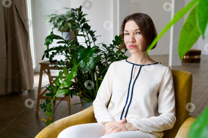 Скачать Сдержанная брюнетка-бизнесвумен сидит в желтом кресле, сложив руки на груди, в современной биофильной гостиной фотосток Ozero