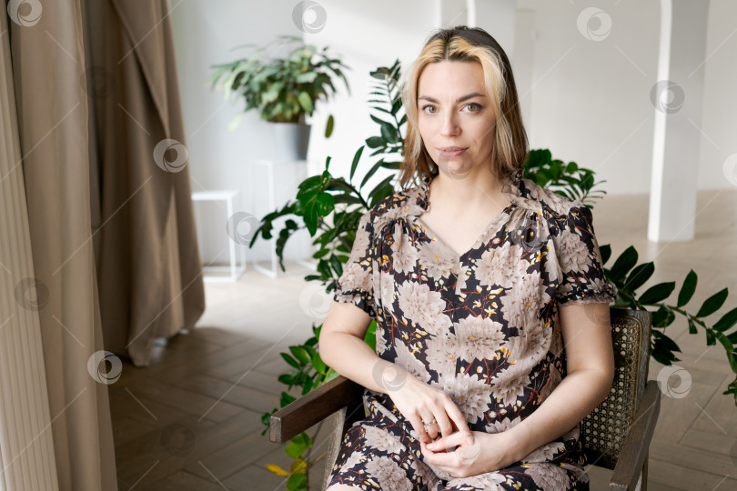 Скачать Женщина в цветочном платье с нежной улыбкой сидит на старинном деревянном стуле в комнате, заполненной растениями фотосток Ozero