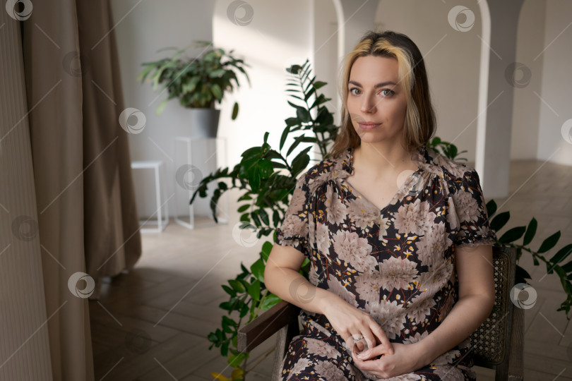 Скачать Безмятежная блондинка в цветочном платье сидит на деревянном стуле в ярком биофильном интерьере фотосток Ozero