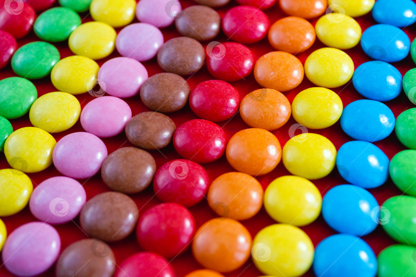 Скачать Радужный узор из круглых цветных шоколадных конфет. Свитс снят крупным планом на красном фоне. Избирательный фокус фотосток Ozero