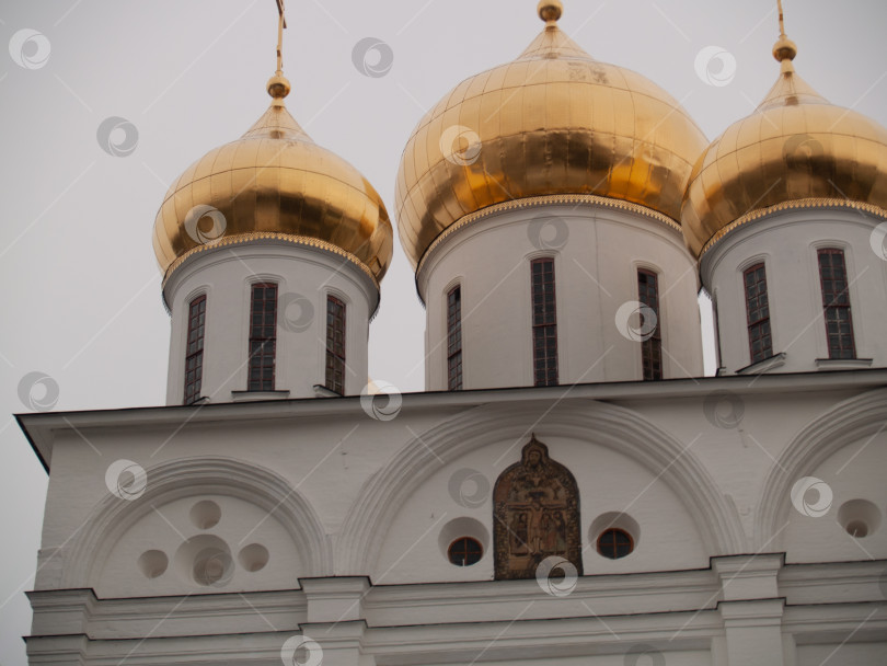 Скачать ДМИТРОВ, РОССИЯ - 11 января 2016 года: Успенский собор в Дмитрове. Часть православного собора с тремя золотыми куполами, белыми стенами и арочными окнами. Икона Христа на фасаде. Традиционный русский архитектурный стиль. фотосток Ozero
