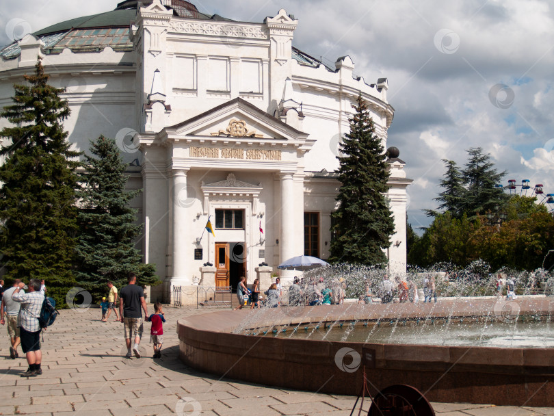 Скачать СЕВАСТОПОЛЬ, РОССИЯ - ИЮЛЬ, 15, 2015: Белоснежное здание музея-панорамы с куполом и колоннами, окружённое зелёными деревьями. На переднем плане — фонтан и гуляющие люди, небо с облаками создаёт живописный фон. фотосток Ozero