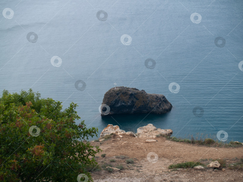 Скачать СЕВАСТОПОЛЬ, РОССИЯ - ИЮЛЬ, 20, 2015: Скала посреди Чёрного моря с крестом на вершине. На переднем плане — каменистый берег и зелёная растительность. Спокойная водная гладь, живописный морской пейзаж Крыма. фотосток Ozero