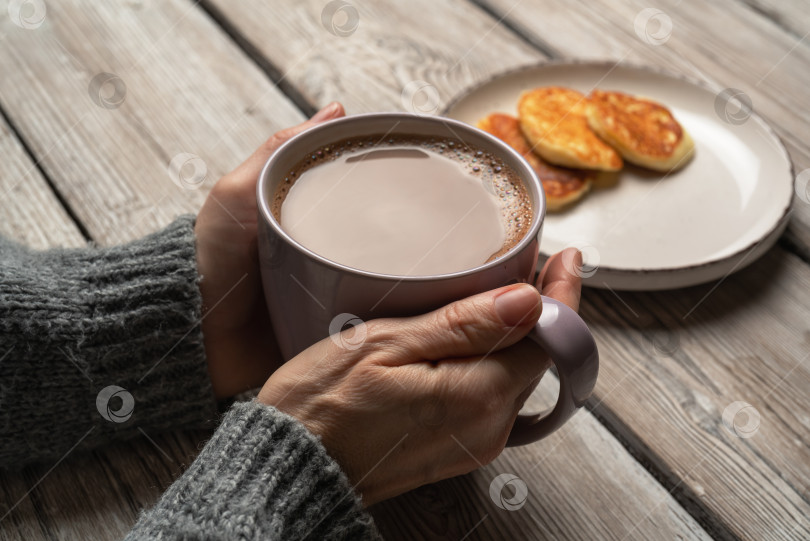 Скачать Женские руки в сером свитере, держащие кружку горячего шоколада рядом с тарелкой блинчиков на деревянном столе в деревенском стиле фотосток Ozero