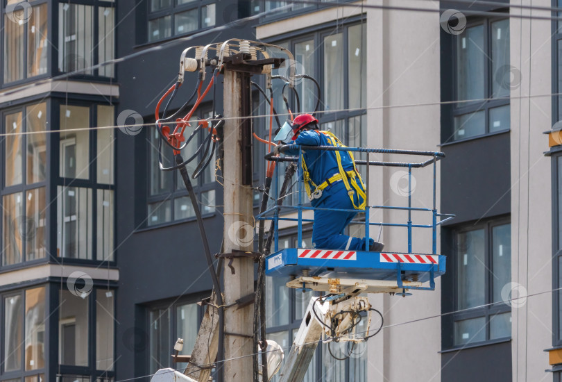 Скачать Электрик, поднятый в люльке башенного подъемника, монтирует провода на столбе на фоне высотного здания фотосток Ozero