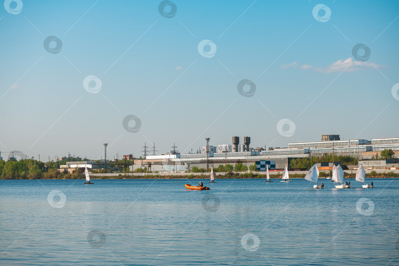 Скачать Екатеринбург, Россия, 08 мая 2026 года. Городской пейзаж с современными зданиями и зелеными деревьями на фоне безмятежного озера и маленькими парусниками, грациозно скользящими по спокойной воде. фотосток Ozero