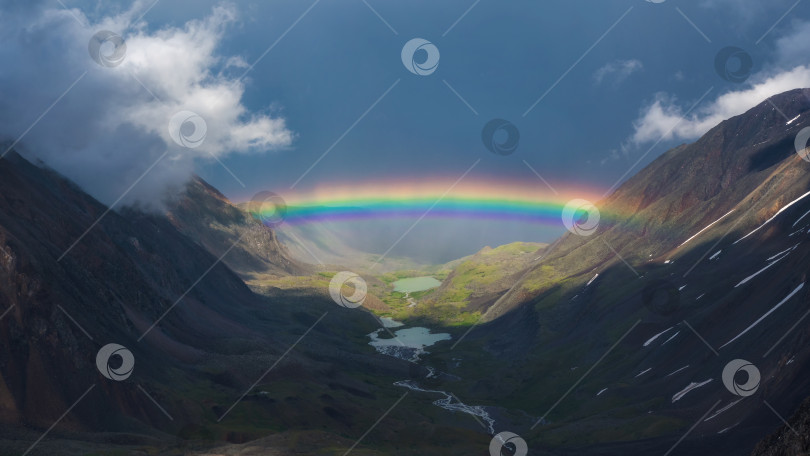 Скачать Полная радуга над горной долиной. Атмосферный альпийский пейзаж со снежными горами и радугой в дождливую и солнечную погоду. фотосток Ozero